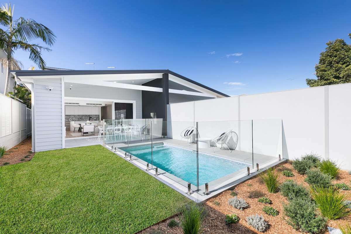 A private outdoor space is depicted, featuring a clear glass fence surrounding a magnesium mineral pool. Lush green grass is present, alongside a landscaped area with various plants. The open-plan living area is visible in the background, allowing for a seamless indoor-outdoor connection.
