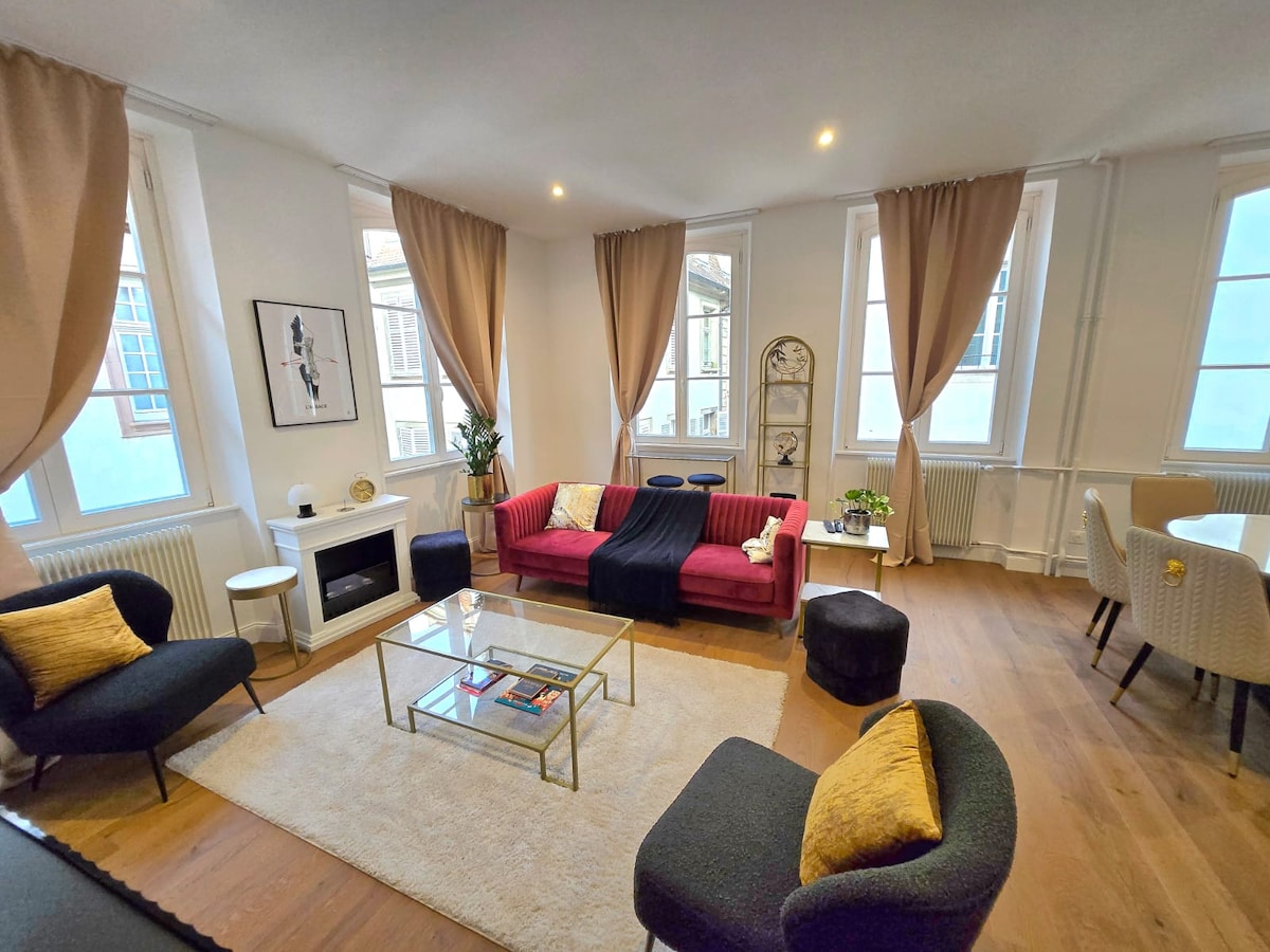 A bright living area is displayed with large windows allowing natural light to fill the space. A red sofa is complemented by two black chairs and a glass coffee table. A cozy rug covers the wooden floor, while decorative plants enhance the ambiance.