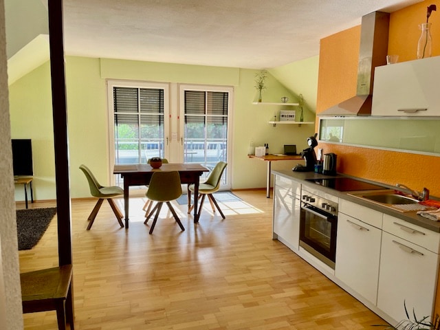 An open living area is highlighted by a dining table surrounded by four chairs, positioned near a large window. A well-equipped kitchen is visible, featuring modern appliances and an orange accent wall. Light wood flooring enhances the spacious feel.