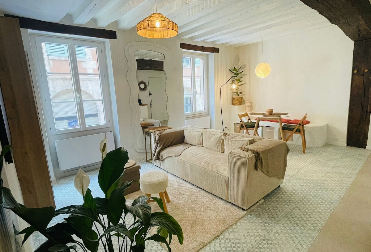 A spacious living area features a light-colored sofa and a round stool placed on a textured rug. A dining table with four chairs is positioned near a large window, providing ample natural light. A decorative mirror and indoor plant add to the space's charm.