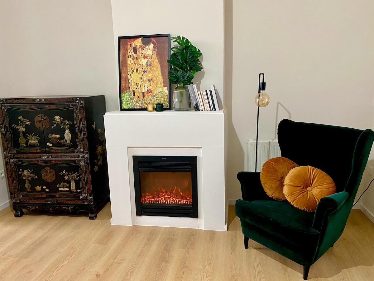 A cozy living area features a modern electric fireplace set within a white mantel. A dark green upholstered armchair with decorative pillows is positioned nearby, alongside an ornate black cabinet displaying decorative objects. Plants and books add a touch of warmth to the space.