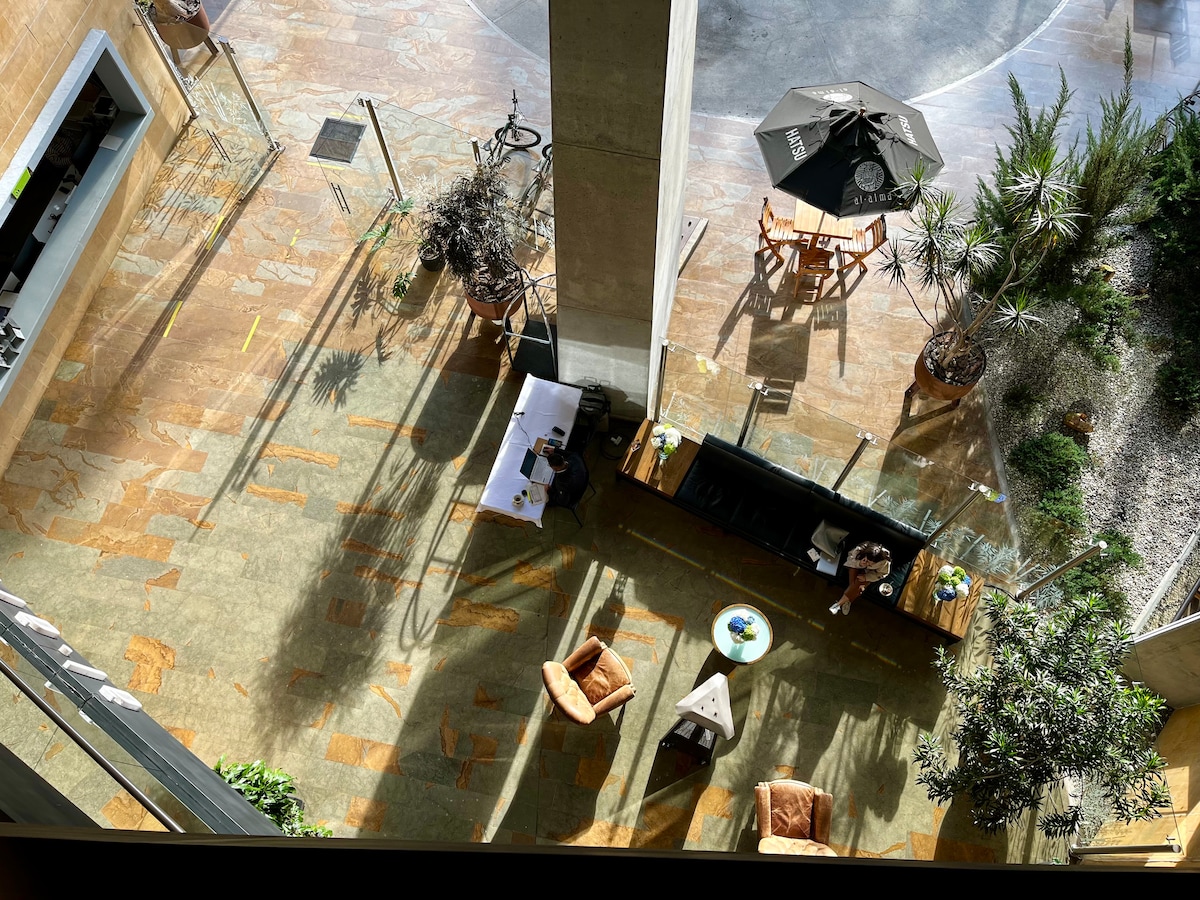 An open outdoor space is shown from an aerial view, featuring stone flooring and various potted plants. A dark umbrella shades a seating area, where wooden tables and chairs are arranged. Lounge chairs are positioned on one side, adding to the inviting environment.