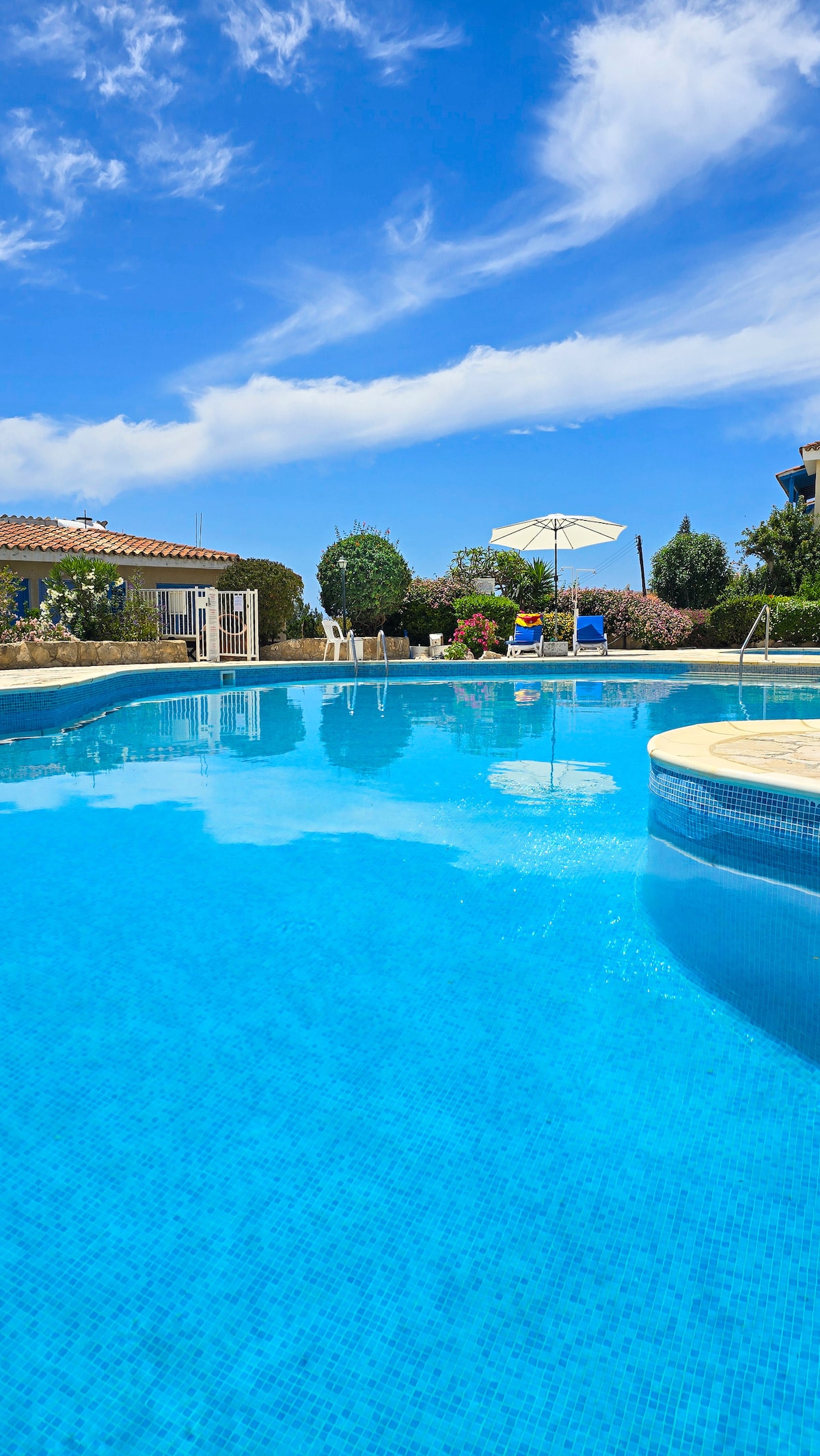 A serene pool area is captured with clear blue water reflecting the sky. Surrounding greenery provides a natural backdrop. A shaded seating area is visible near the edge, accompanied by lounge chairs and an umbrella for relaxation.