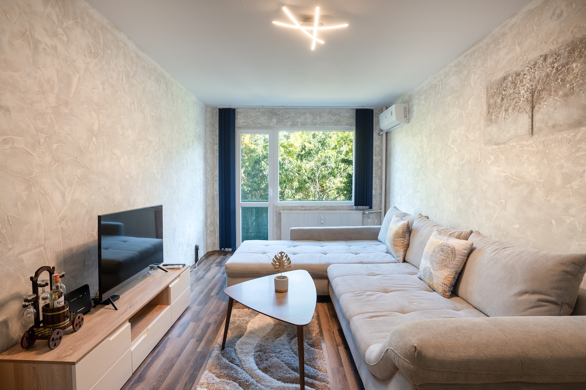 A spacious living room is highlighted by a large sectional sofa and a modern coffee table, positioned in front of a flat-screen TV. Natural light filters through the large window adorned with blue curtains, while decorative artwork adds a subtle touch to the textured walls.