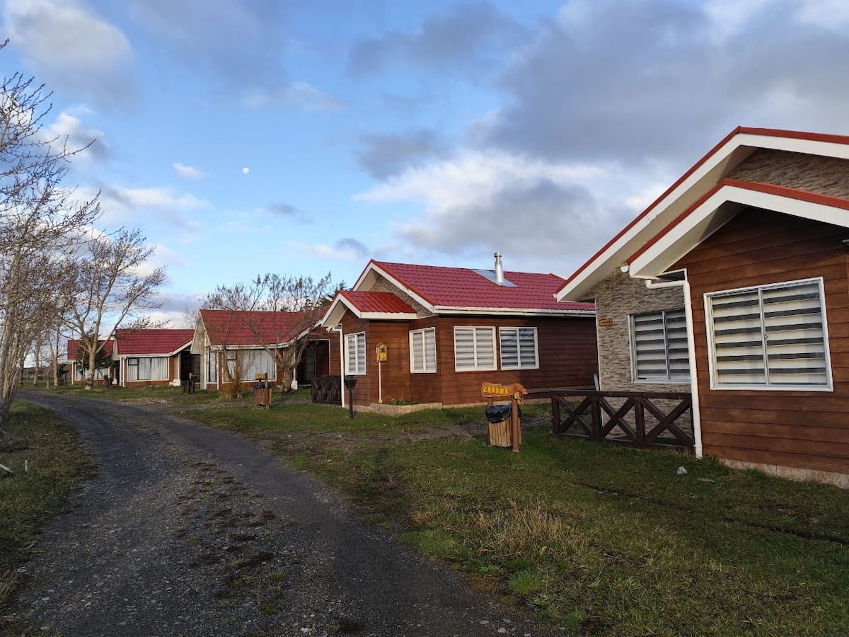 A row of wooden cabins with red roofs is visible along a gravel path. Each cabin features multiple windows with horizontal shutters, and greenery is present on the property. A clear sky with scattered clouds complements the serene landscape.