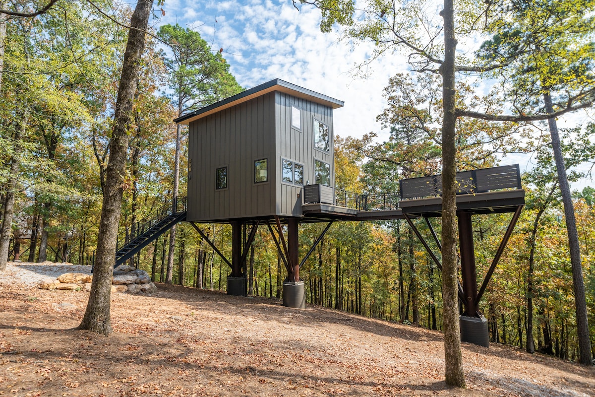 A modern treehouse is situated on elevated stilts amidst autumn foliage. Large windows allow natural light to fill the interior, while a spacious deck with seating invites outdoor enjoyment. Surrounding trees display vibrant colors, contributing to the serene natural setting.