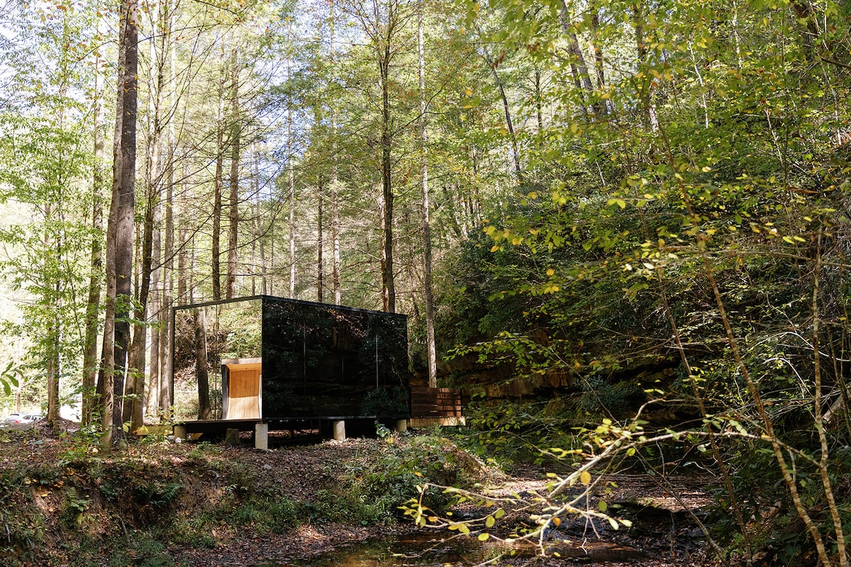 A modern glass cabin is situated amid lush green trees, reflecting the surrounding landscape. The cabin features a mirrored exterior, creating a harmonious blend with nature. A small creek runs beside it, adding to the serene woodland setting.