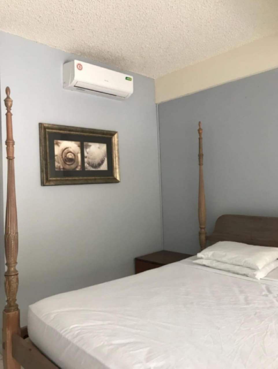 A bedroom features a simple wooden bed with a crisp white sleeping surface and two pillows. An air conditioning unit is mounted above the bed, while a framed artwork of shells adorns the wall, adding a touch of design to the space.