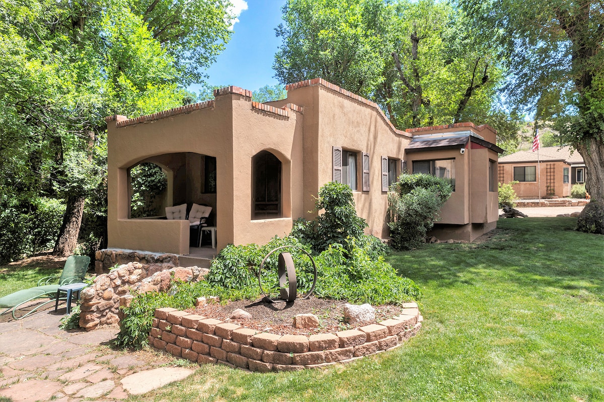 The exterior of a rustic adobe cottage is featured, showcasing a shaded porch with seating. Lush greenery surrounds the property, and a neat stone border defines the garden area. The soft sunlight illuminates the peaceful yard, creating a calm outdoor setting.