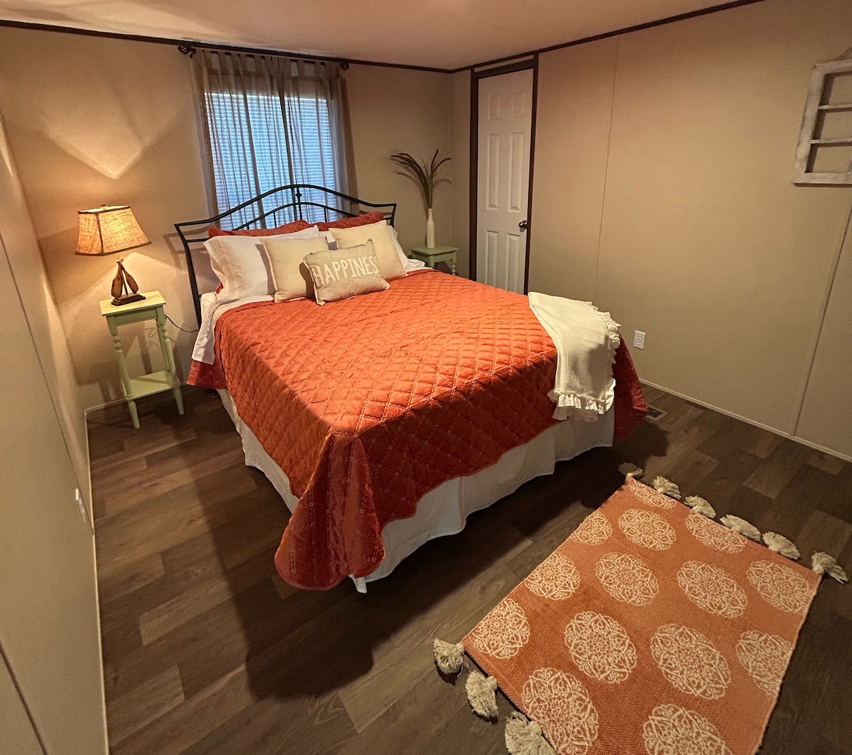 A welcoming bedroom is presented with a large bed layered in an orange quilt and white linens. A decorative pillow is placed at the head of the bed. Soft lighting from a bedside lamp creates warmth, and a patterned area rug complements the wooden flooring.