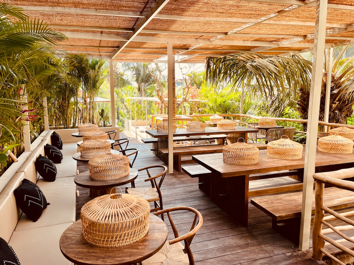 An outdoor dining area is featured, showcasing wooden tables surrounded by natural greenery. Light woven baskets sit atop the tables, while a canopy provides shade. Various seating options include chairs with a minimalist design, creating a relaxed atmosphere for guests to enjoy.
