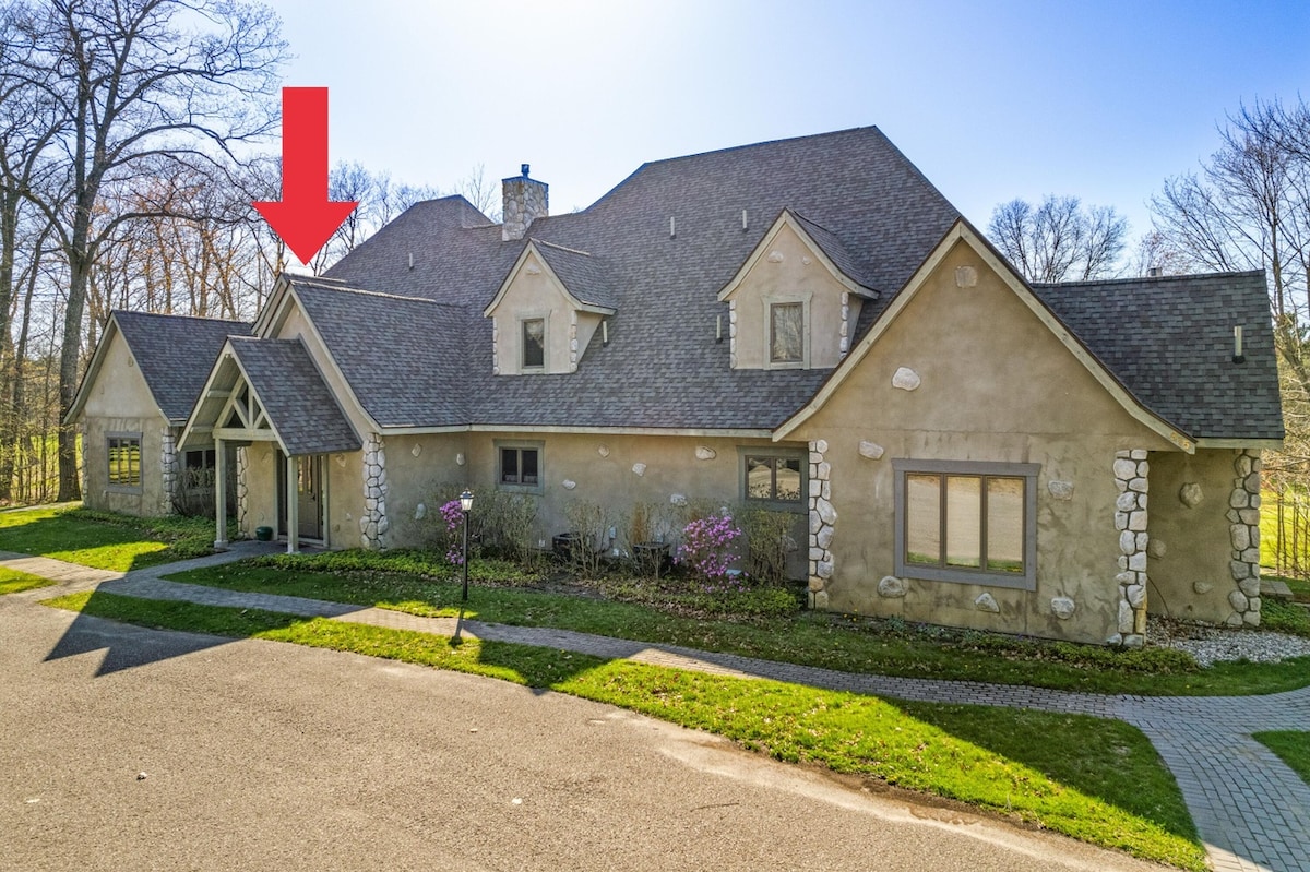 A charming French villa is depicted with a textured exterior featuring a blend of stone and siding. The entrance is highlighted by a small covered porch, and large windows allow for ample natural light. Surrounding greenery adds to the serene setting.