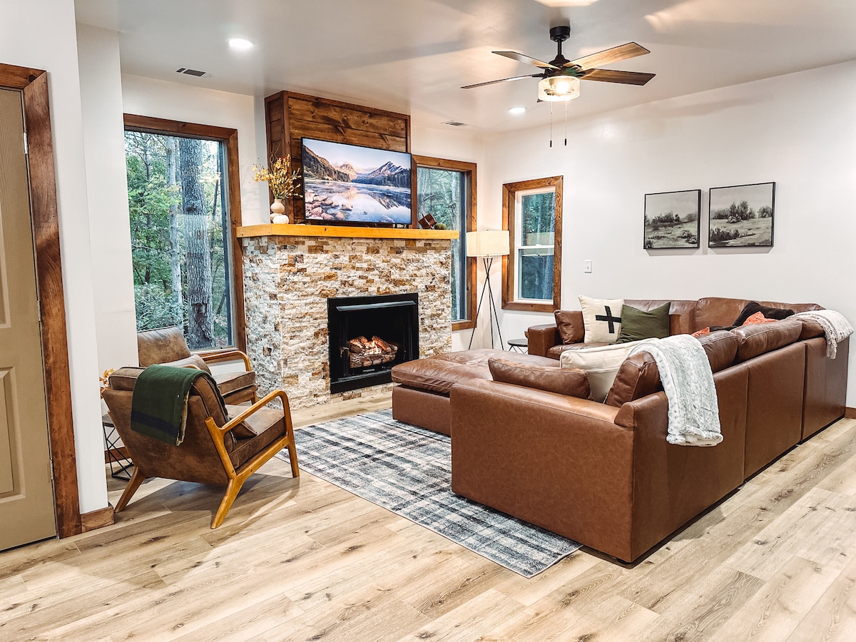 The spacious living room features a large sectional sofa in warm brown tones, complemented by a stone fireplace and a mounted television. Tall windows provide views of the surrounding trees. A cozy area rug adds texture, and soft furnishings enhance the comfortable ambiance.