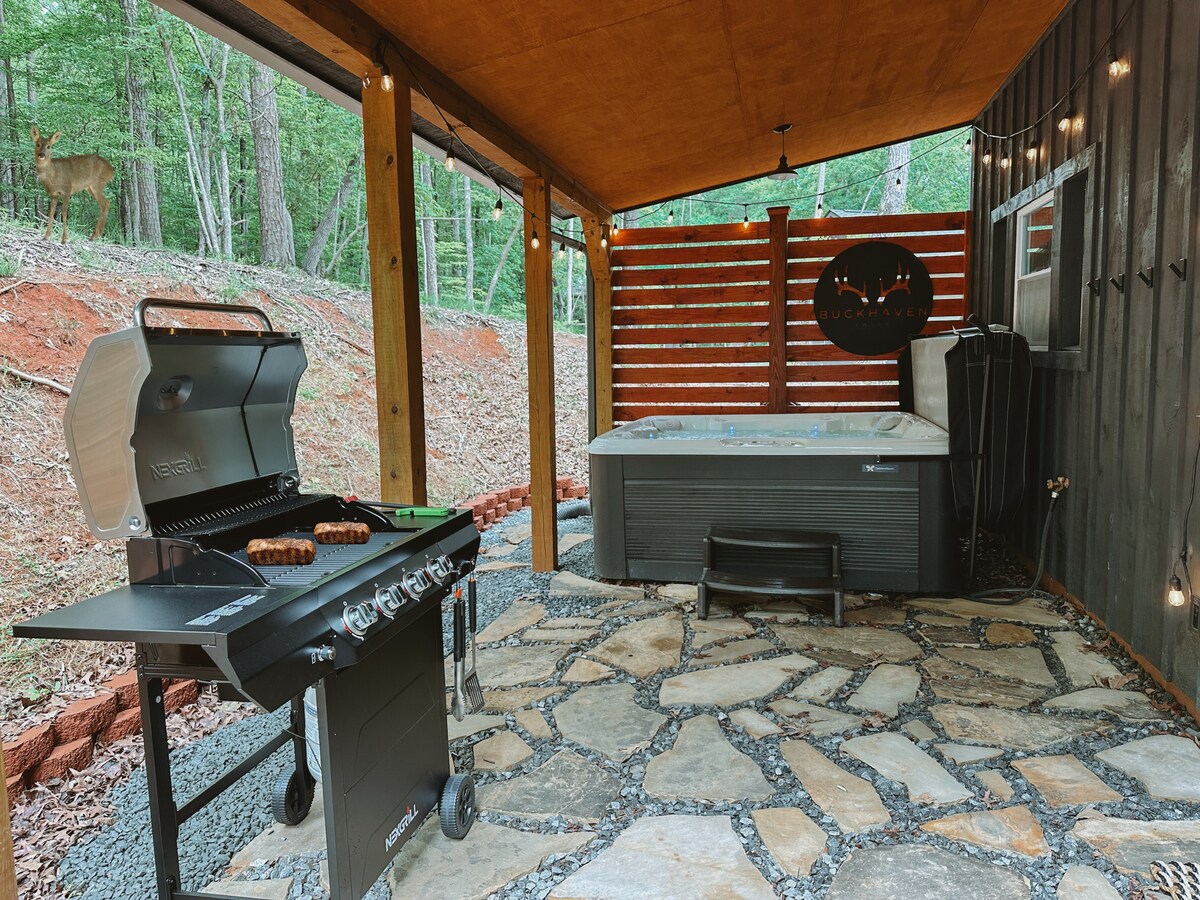 An outdoor space features a grill beside a hot tub on a stone patio, surrounded by trees. Soft lighting adds a cozy touch, and a wooden privacy screen offers seclusion. The nearby lodge sign is visible, enhancing the setting's rustic appeal.
