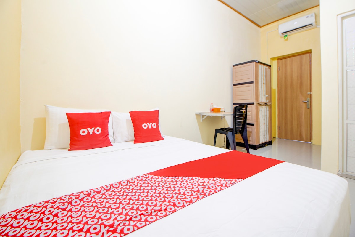 A clean and modern room is shown featuring a spacious bed with white linens and vibrant red accent pillows. A simple desk and chair are positioned against the wall, providing a workspace. A wardrobe stands in the corner, and light-colored walls enhance the brightness.