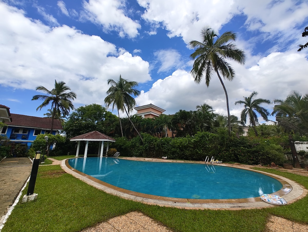 A large swimming pool surrounded by lush greenery is highlighted under a partially cloudy sky. Palm trees provide shade around the pool area. A gazebo sits nearby, offering a relaxing spot to enjoy the views.