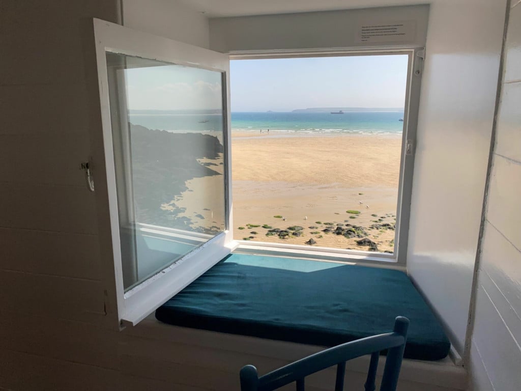 A window seat with a deep blue cushion overlooks a sandy beach and calm turquoise waters. The window opens wide, providing unobstructed views of the shoreline and distant ships, creating a bright and airy atmosphere within the room.