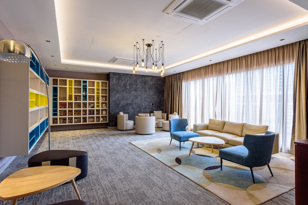 A spacious lounge area features large windows allowing natural light to fill the room. Comfortable seating, including plush sofas and armchairs, surrounds a coffee table. A decorative bookshelf is visible, filled with a variety of books, adding a touch of style to the space.