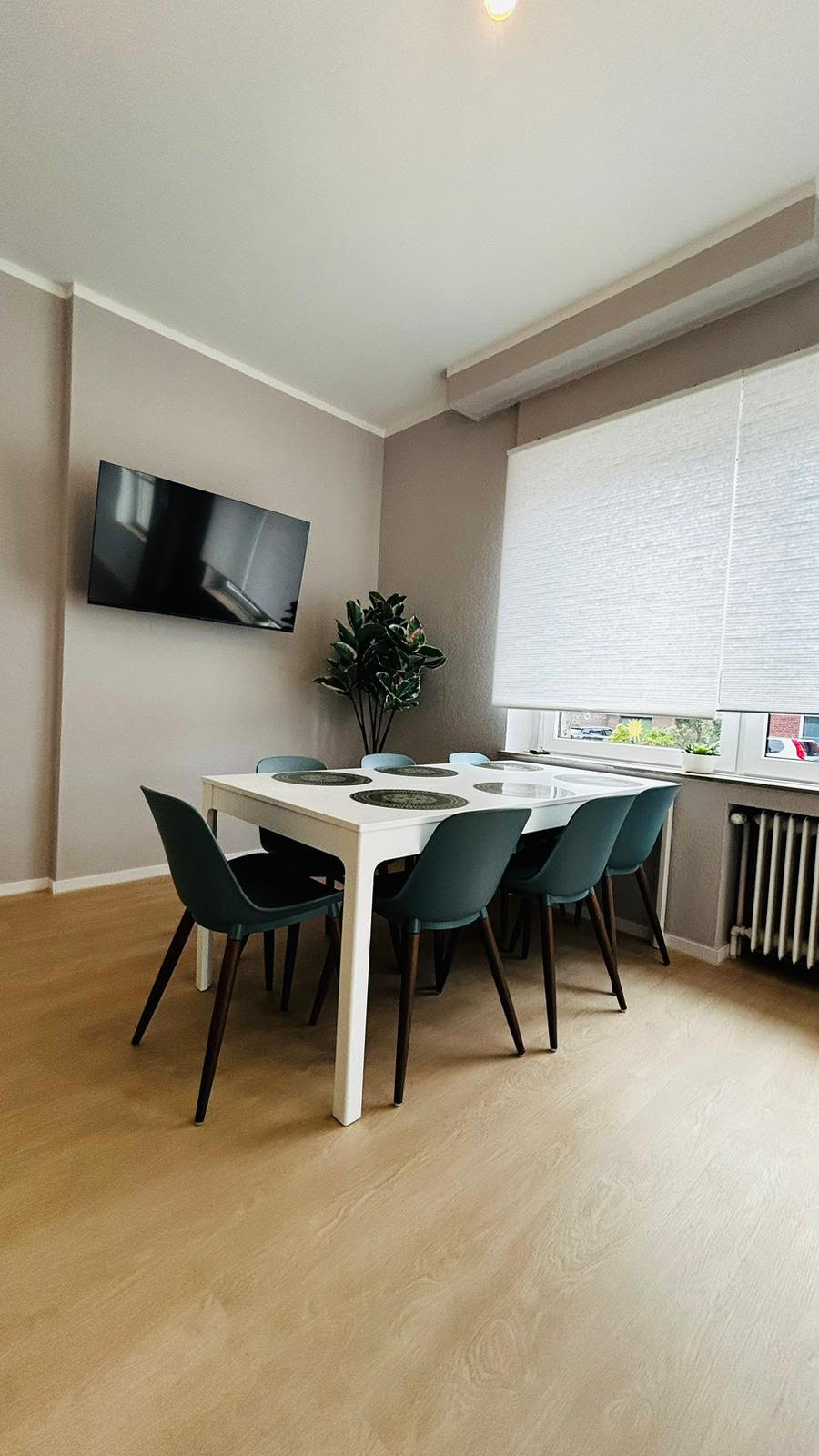 A dining area is presented with a white table surrounded by six teal chairs. A large window allows natural light to fill the space, adorned with a light-colored roller shade. A flat-screen TV is mounted on the wall, while a potted plant adds a touch of greenery.