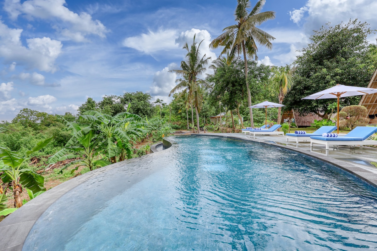 A tranquil infinity pool extends invitingly, surrounded by lush greenery and palm trees. Lounge chairs with umbrellas are positioned nearby, offering a calm spot for relaxation. The clear blue sky enhances the serene atmosphere, inviting guests to unwind in nature.