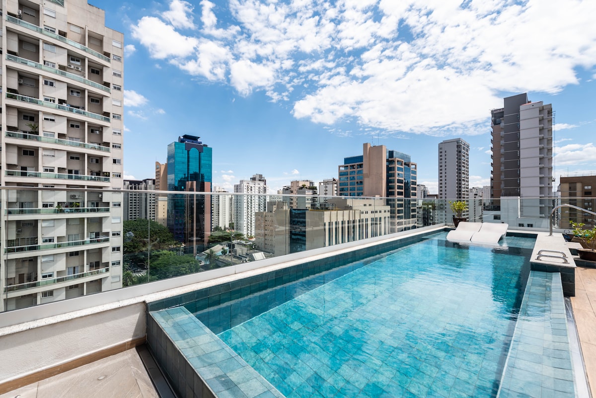 A rooftop pool is showcased, presenting a clear view of the surrounding skyline with modern buildings. The pool features reflective blue tiles and two lounge chairs positioned on the edge. Bright clouds are visible in a partially sunny sky.