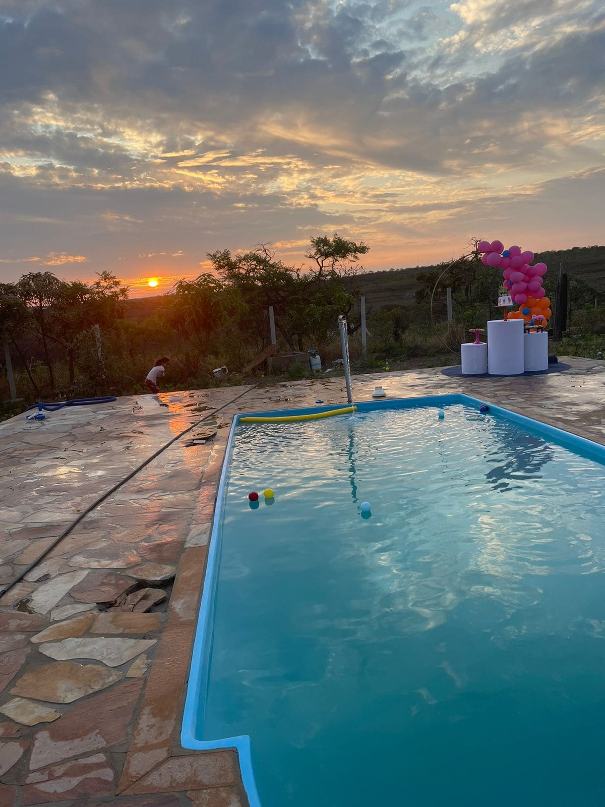 A tranquil swimming pool is bordered by a stone patio, reflecting the warm colors of the sunset. In the background, gentle hills and greenery provide a natural setting. Colorful decorations are arranged nearby, enhancing the festive atmosphere.