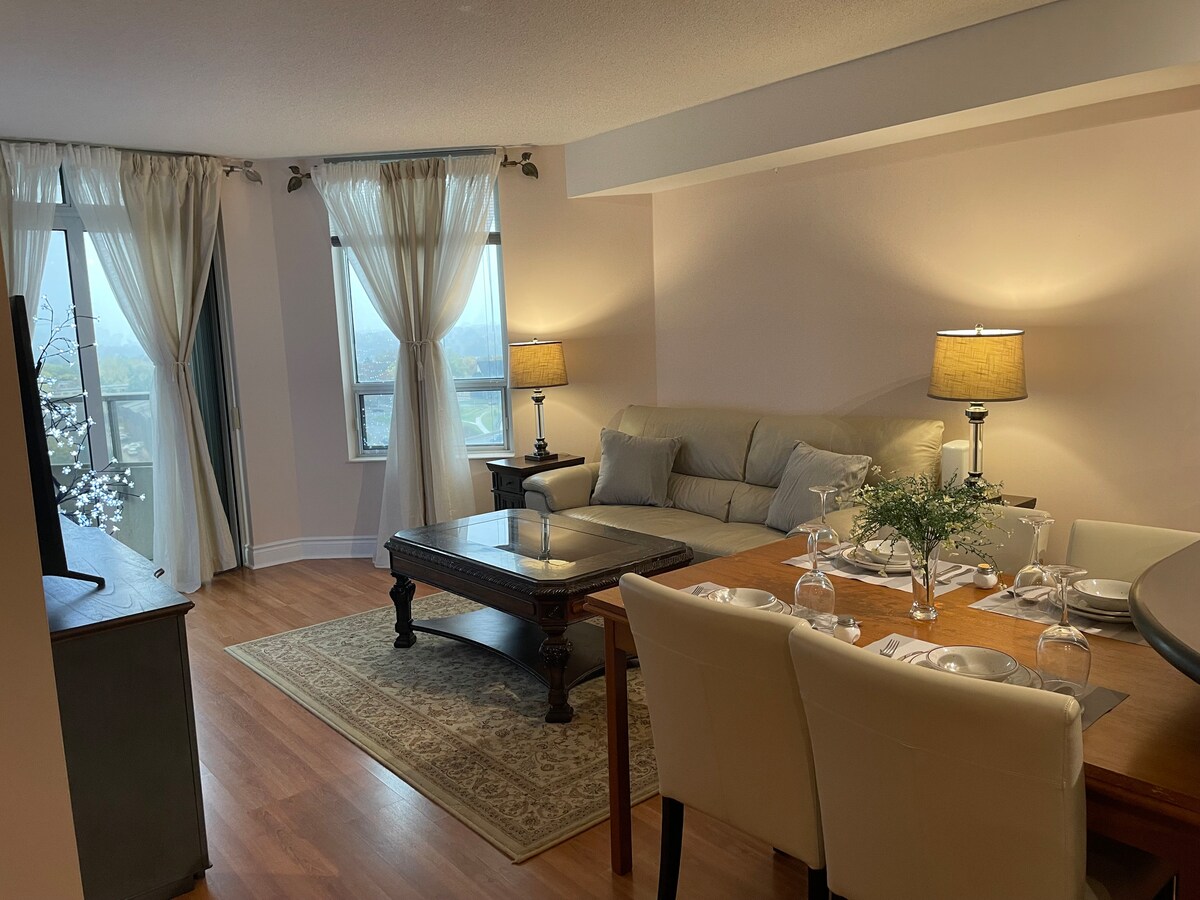 A spacious living area is presented with light wood flooring and neutral-colored walls. A comfortable sofa is positioned next to a coffee table, while a dining table is set with glassware and simple decor. Natural light enters through large windows, adorned with sheer curtains.