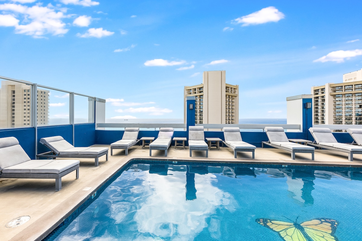 The rooftop pool area is highlighted by a clear blue sky with scattered clouds. Several lounge chairs are arranged around the pool, reflecting in the water. Tall buildings in the distance provide a backdrop to the serene setting.