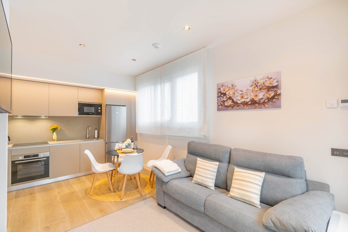 A modern living space features a comfortable gray sofa accompanied by decorative pillows. A small dining table is set for two, and a kitchen area with sleek cabinetry and appliances, including a microwave and refrigerator, is visible. Natural light filters through sheer curtains.