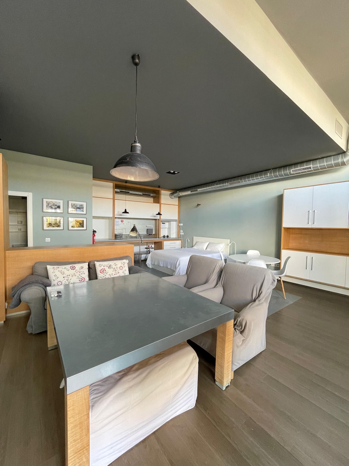 A spacious loft apartment is visible, featuring a modern design with a gray ceiling. A dining table is set in the foreground, complemented by soft armchairs. In the background, a comfortable sleeping area with a large bed and additional furniture creates a functional living space.