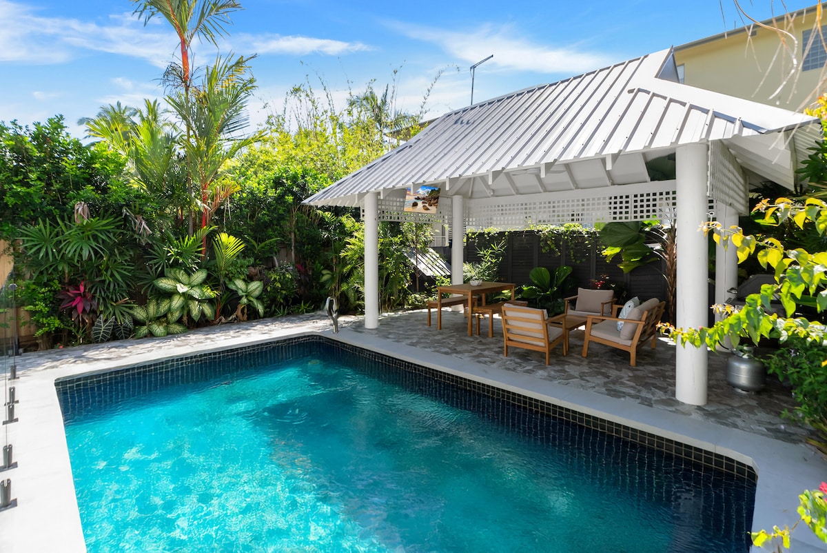 A bright outdoor space features a serene pool surrounded by lush tropical foliage. A shaded cabana offers a dining area alongside comfortable seating, presenting a perfect spot for relaxation. Sunlight filters through the leaves, creating a calming atmosphere in this private retreat.