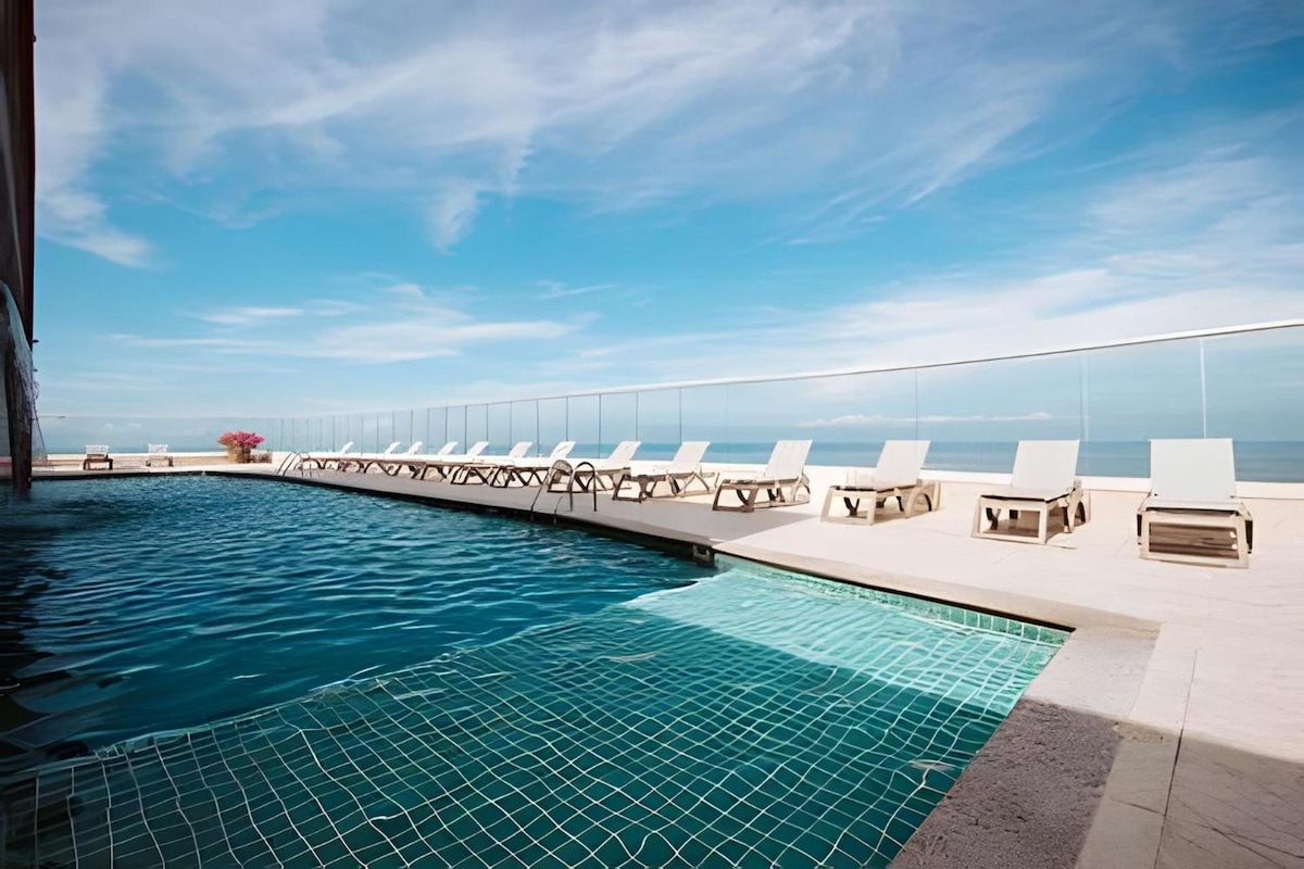 A rooftop pool area features an infinity edge that overlooks the ocean. A series of lounge chairs are arranged along the deck, with a modern design complementing the serene atmosphere. Bright blue skies enhance the tranquil setting, providing a perfect spot for relaxation.
