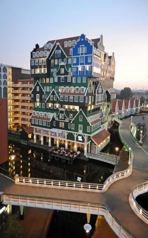A uniquely designed multi-story building is depicted, featuring a mix of colorful façades in green, blue, and red. The structure reflects traditional Dutch architecture, while the surrounding waterway enhances the visual interest. Soft evening light casts a warm glow on the scene.