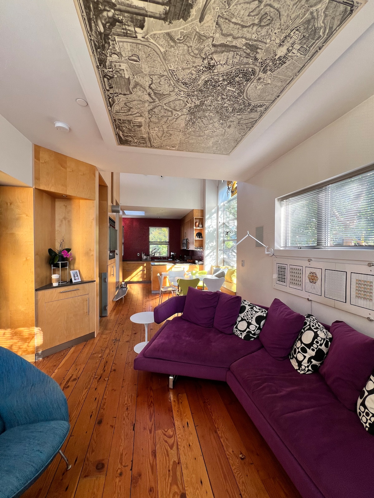 An open-concept living area features a deep purple sectional sofa adorned with black and white patterned cushions. A wooden floor adds warmth to the space, while large windows allow natural light to illuminate the room. A vintage map hangs from the ceiling, enhancing the artistic ambiance.