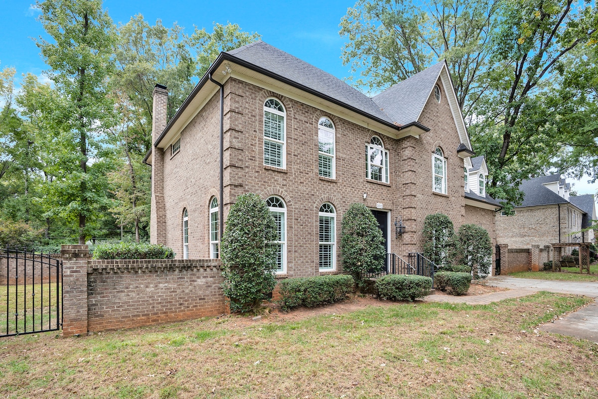 The exterior of a traditional three-level brick house is depicted, featuring a well-maintained lawn with manicured shrubbery along the front. Large arched windows allow natural light to illuminate the interior. A paved driveway leads to the entrance, surrounded by a gated border.