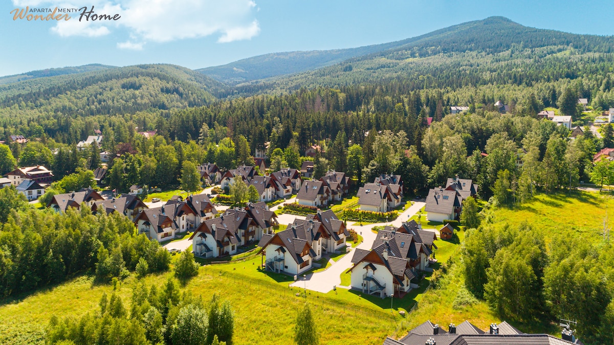 An aerial view captures a tranquil residential area surrounded by lush greenery and hills. The layout features multiple two-story cabins with sloped roofs, nestled along winding paths, offering a sense of community amid scenic natural surroundings.