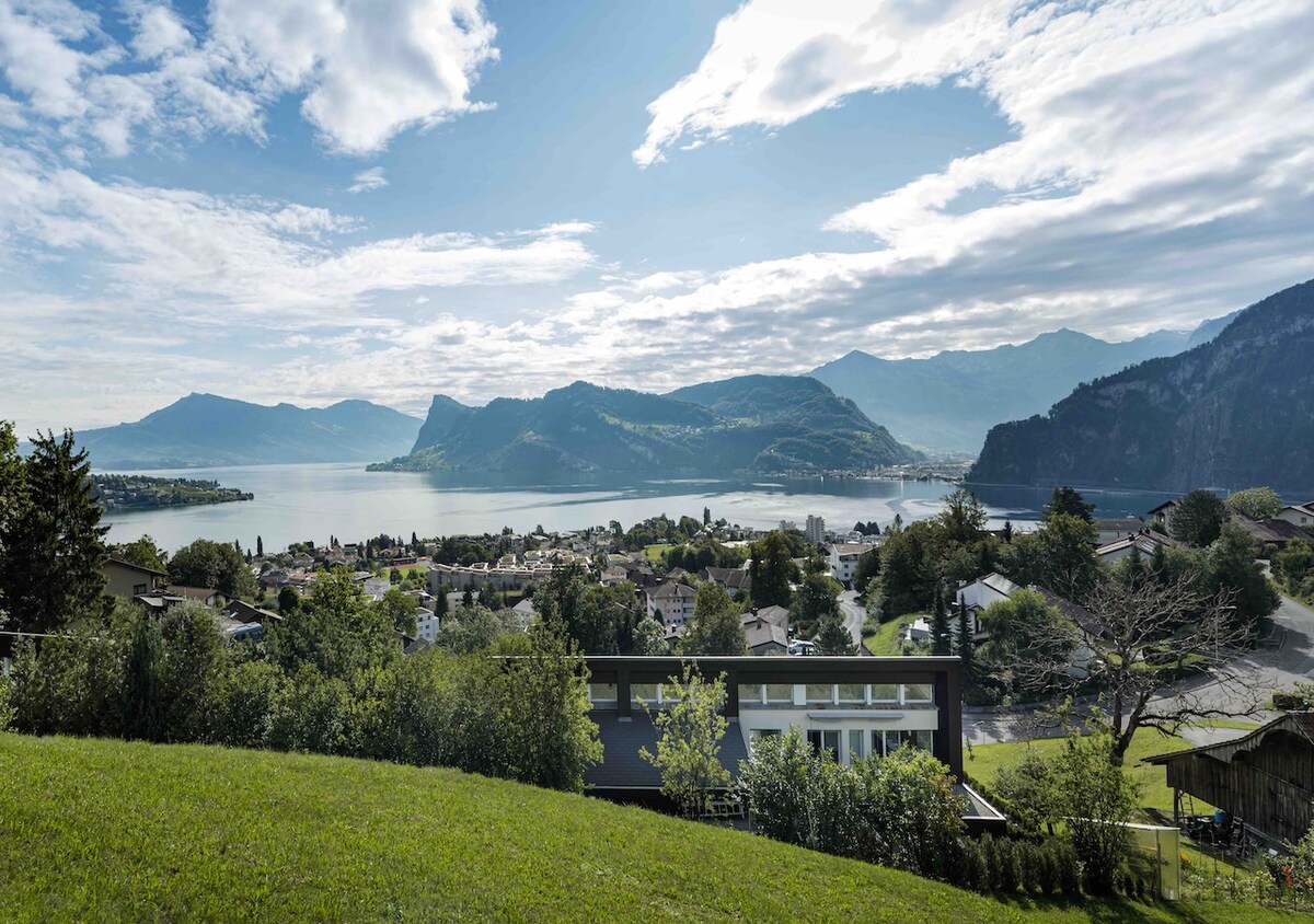 A panoramic view captures the serene beauty of Lake Lucerne and surrounding mountains. The horizon is framed by rolling hills and lush greenery, while the village below is nestled along the lakeshore. Soft clouds drift across a bright sky, enhancing the tranquil atmosphere.