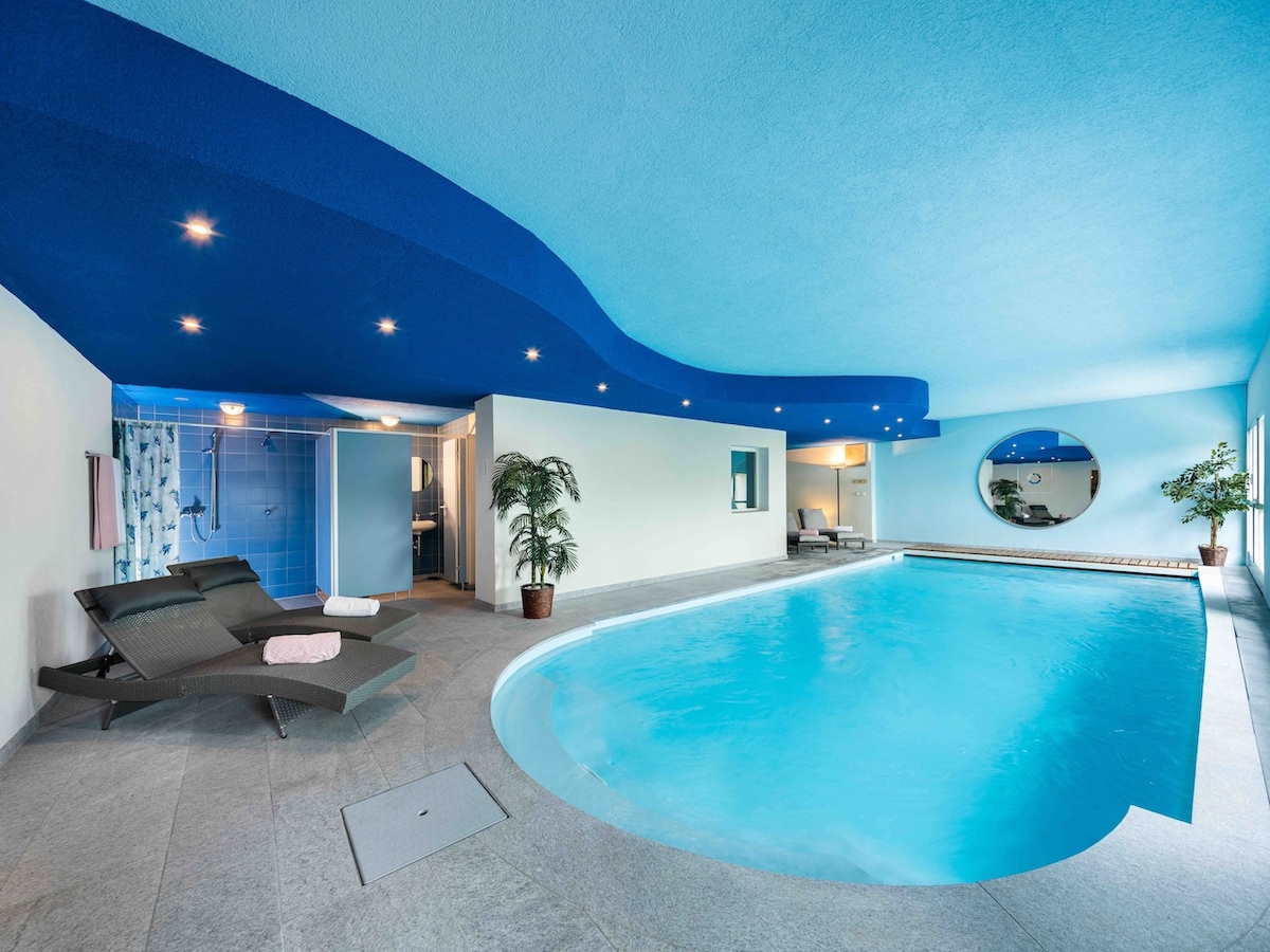 An indoor heated swimming pool is surrounded by sleek tiled flooring and walls painted in a calming blue. Lounge chairs are positioned beside the pool, and a potted plant adds a touch of greenery. A shower area is visible, with the pool reflecting the soft overhead lighting.
