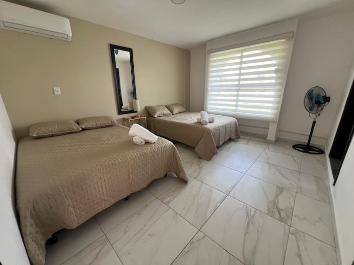 Amplia y luminosa habitación equipada con cama Queen y matrimonial, TV, ventilador y gran ventanal. Ideal para descansar en un ambiente fresco y cómodo después de disfrutar la playa.