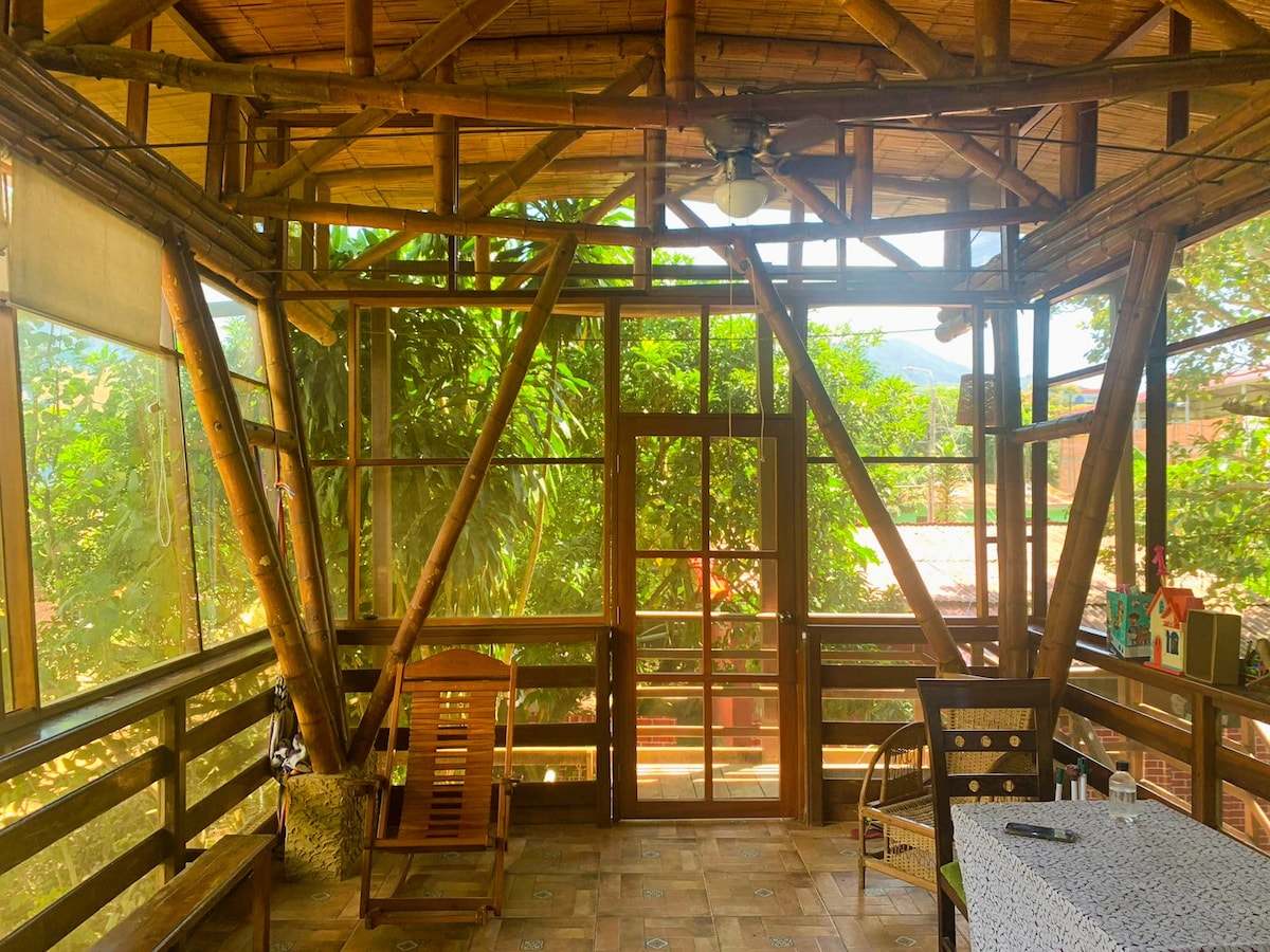 A terrace area features a bamboo structure with large windows that invite abundant natural light. The space includes traditional rocking chairs and a table with a tablecloth. Lush greenery is visible through the windows, creating a peaceful atmosphere connected to the outdoors.