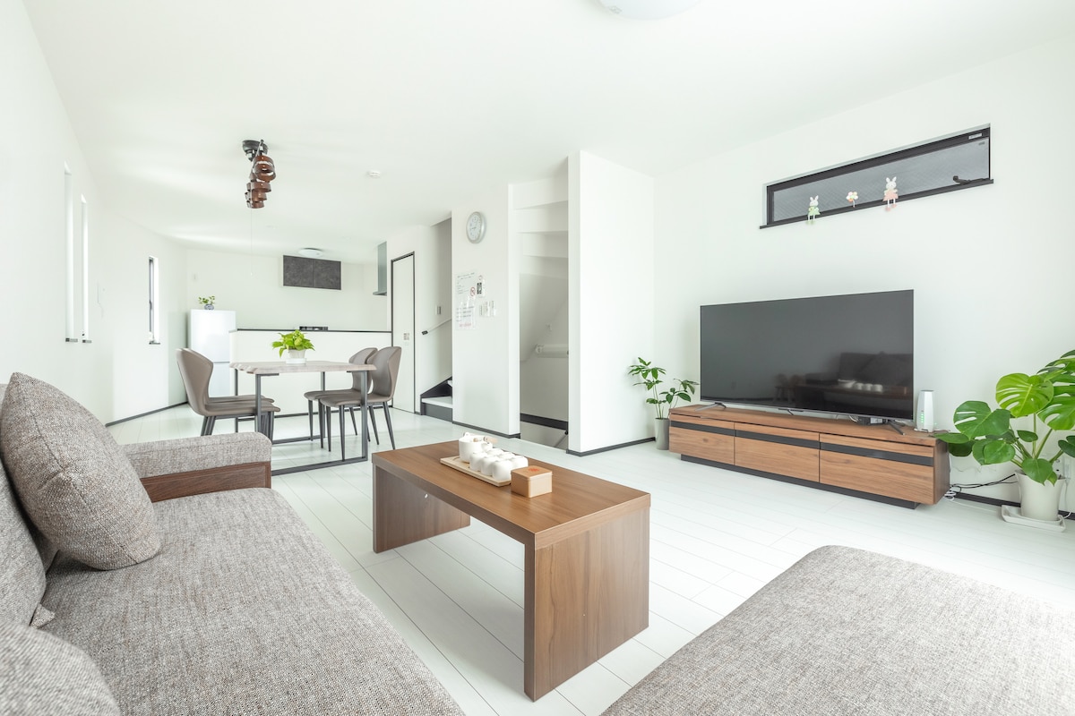 A bright and airy living space is shown with light-colored walls and modern furnishings. A comfortable sofa faces a large television on a wooden stand. A dining table for six is positioned nearby, and several indoor plants add a touch of greenery to the room.
