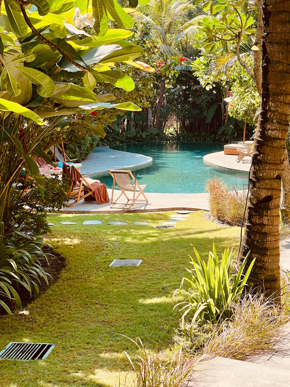 A lush garden path leads to a serene swimming pool surrounded by tropical foliage. Sunlight filters through the greenery, highlighting the soft curves of the pool's edges. A few lounge chairs are positioned nearby, inviting relaxation amid the natural beauty.
