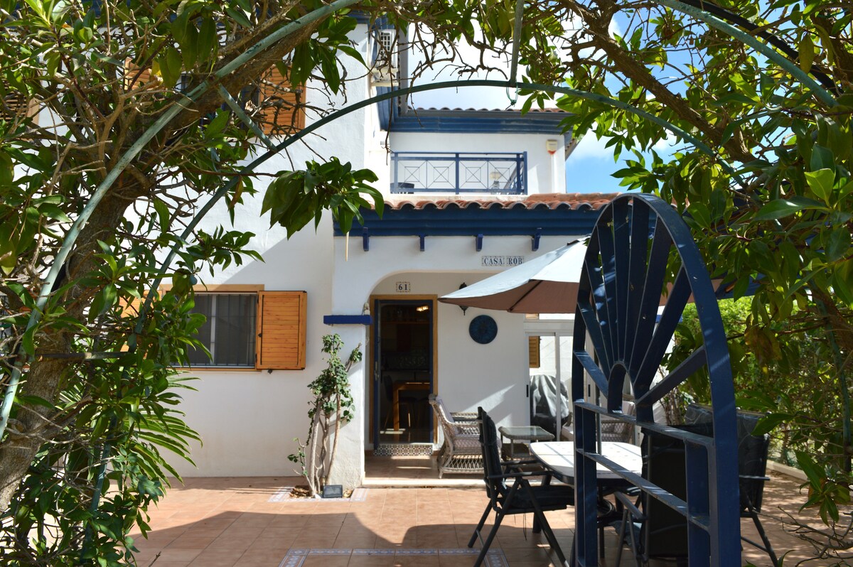 A charming entrance is framed by lush greenery, leading to a two-story home with warm wooden shutters. A shaded outdoor dining area features a table and chairs, inviting guests to enjoy meals al fresco. The entrance is marked by a decorative blue archway.