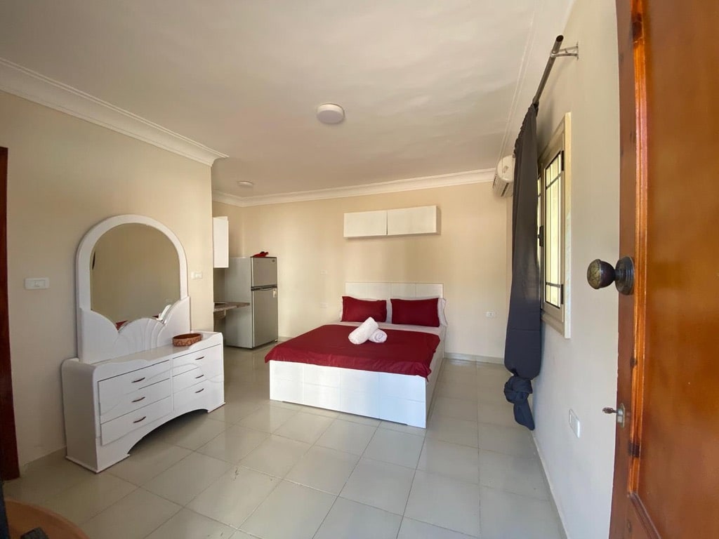 A neutral-toned bedroom is equipped with a spacious bed dressed in red linens. A white dresser and overhead shelving provide storage, while a small refrigerator is visible in the background. Natural light enters through a window, enhancing the room's simplicity.