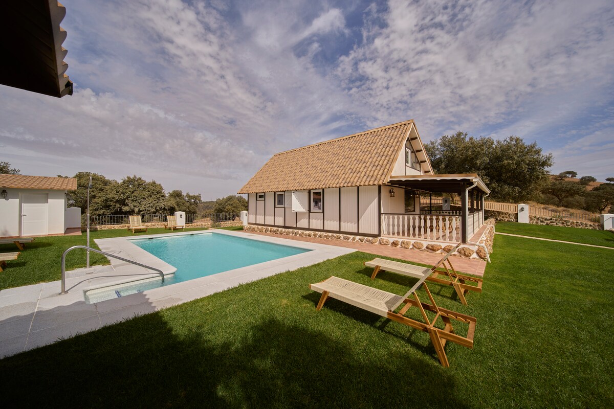 A charming wooden house is situated adjacent to a private swimming pool. Sun loungers are arranged on the manicured lawn, offering a relaxing spot. The structure's architecture features a tiled roof and a spacious front porch, surrounded by lush greenery and a serene landscape.