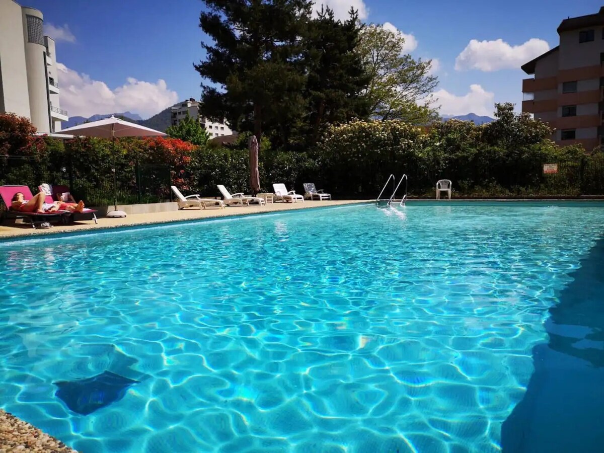 A shared swimming pool is surrounded by lounge chairs, offering a tranquil space for relaxation. The water is clear and reflects the blue sky with fluffy clouds. Lush greenery borders the pool area, contributing to the overall serene environment.