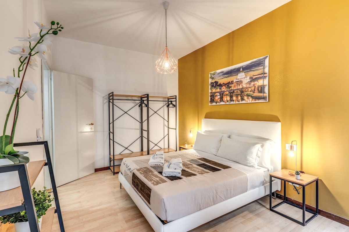 A bright bedroom features a large bed with neutral bedding and decorative pillows. A vibrant yellow wall complements artwork above the bed, depicting a cityscape. Minimalist shelving units are positioned in the room, while a door leads to an adjoining space. Natural light fills the area.