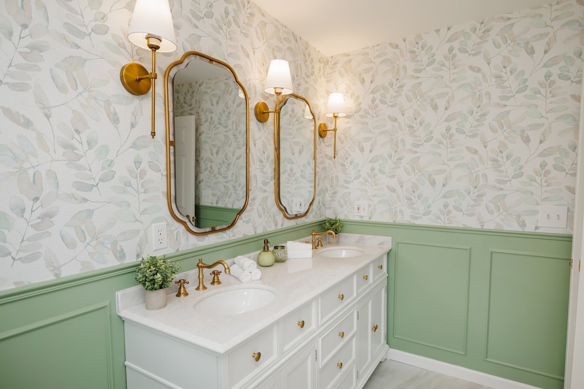 A modern bathroom features dual sinks framed by elegant mirrors. Soft-hued wallpaper with botanical patterns complements green wainscoting. Warm lighting is provided by wall-mounted sconces, creating a calming atmosphere. Decorative plants add a touch of greenery to the space.