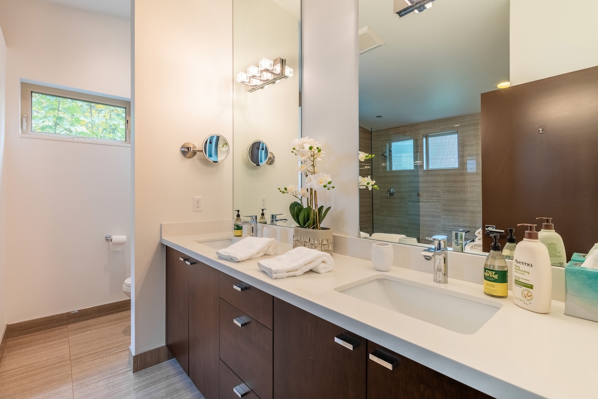 A modern bathroom is depicted, featuring a long vanity with a white countertop and dark wood cabinetry. A large mirror reflects an array of toiletries, while soft lighting enhances the space. A small window allows natural light to enter, complementing the neutral tiles.