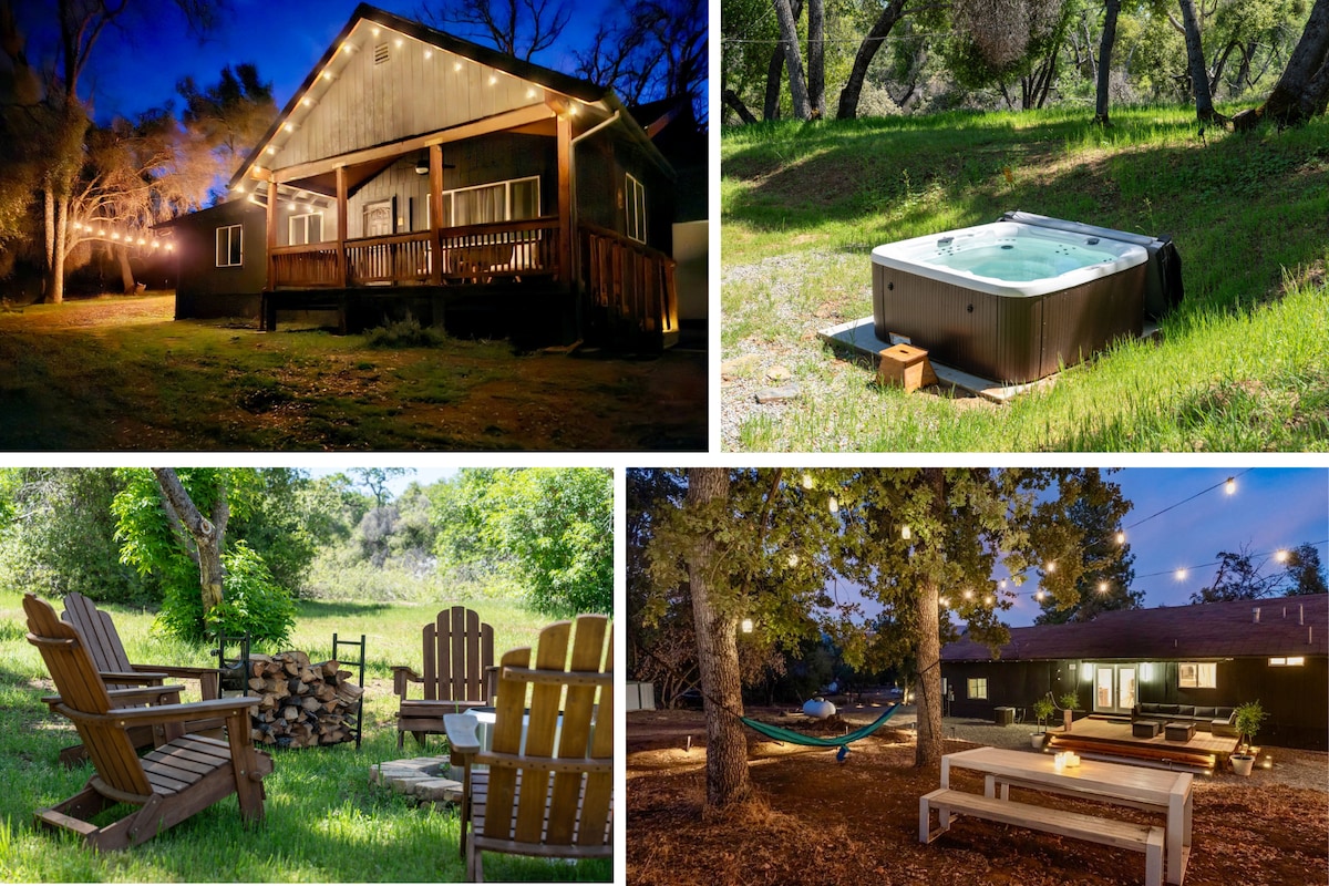 An inviting cabin is illuminated by warm lights, surrounded by natural greenery. A hot tub is positioned nearby, nestled in the grass. Outdoor seating arrangements include comfortable chairs, and a fire pit is surrounded by rustic wooden chairs, providing a cozy gathering space.